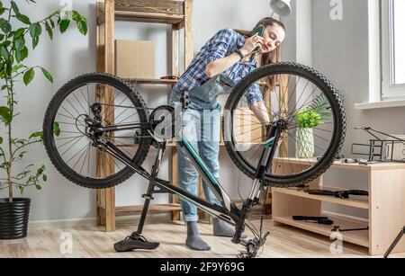 A young woman is fixing a bicycle and using a mobile phone. Concept of maintenance and preparation of the bike for the new season Stock Photo
