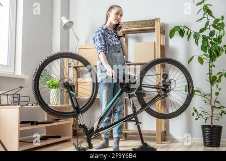 A young woman is fixing a bicycle and using a mobile phone. Concept of maintenance and preparation of the bike for the new season Stock Photo