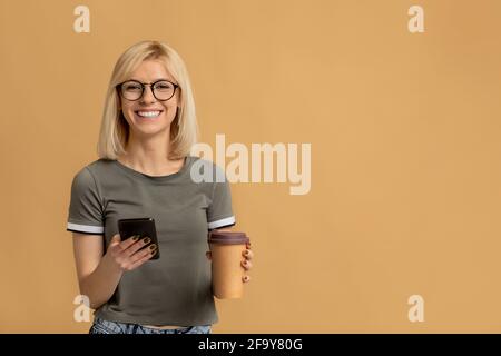 Modern student holding takeaway coffee and typing on smartphone on beige background with empty space Stock Photo