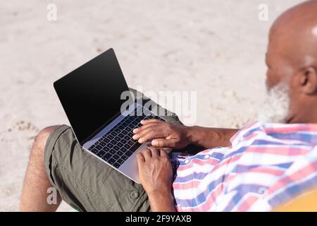 Senior african american man using laptop while sitting on deck chair at the beach Stock Photo