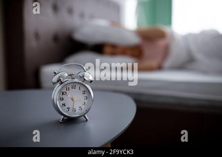 Alarm clock on bedside table and blurred view of young woman unwilling to wake up, covering ears with pillow in bed Stock Photo