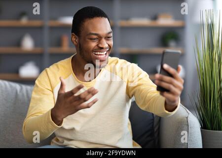 Joyful black man having video conversation on phone, home interior Stock Photo