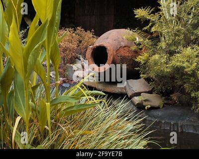 An old shabby earthenware jug on side by a pond among green plants. Landscaping. Resting place Stock Photo