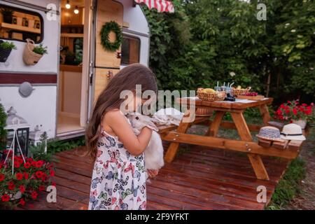 Portrait of a little girl in a straw hat sits near a trailer truck at home on a wooden floor, holding a fluffy rabbit in her hands. Laughs, plays with Stock Photo
