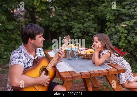 Young brunette father is playing songs guitar for little beautiful daughter. Man and girl are sitting at wooden table in the backyard enjoying the mus Stock Photo
