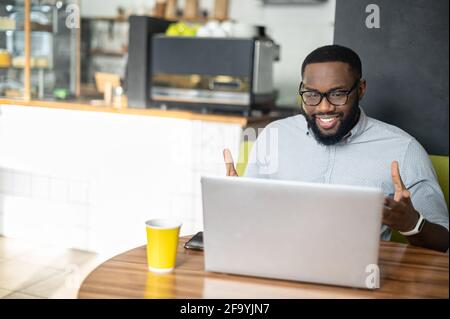 Smiling African-American guy is using a laptop for video call sitting in the cozy cafe. A multiracial young man talks online, has video meeting working remotely Stock Photo