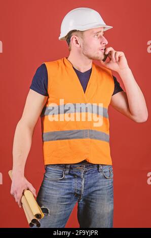 Man, foreman in helmet speaking on phone, red background. Architect calling engineer to fix plan. Engineer, architect, on busy face speaks on Stock Photo