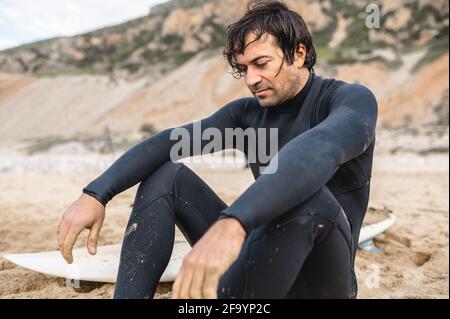 Portrait of young man wearing diving suit sitting on the sand in the beach. Exhausted male resting after surfing. Stock Photo