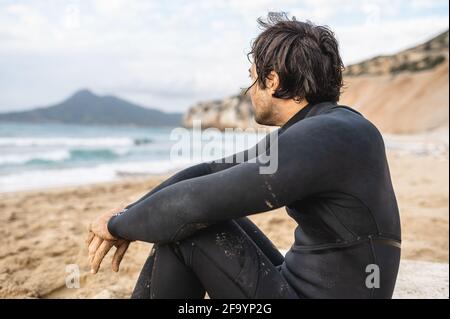 Back view young male wearing diving suit sitting on the sand and looking at the sea. Wet man resting after swimming. Stock Photo