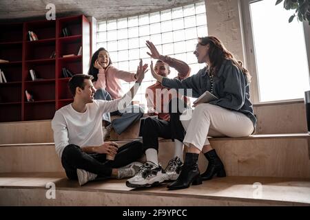 happy interracial students smiling while giving high five and sitting on stairs, senior 2021 Stock Photo