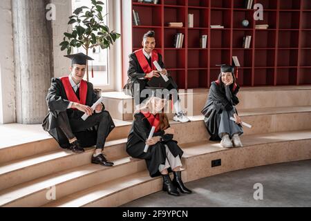 happy interracial students in gowns and caps holding diploma and sitting on stairs, graduation class 2021 Stock Photo