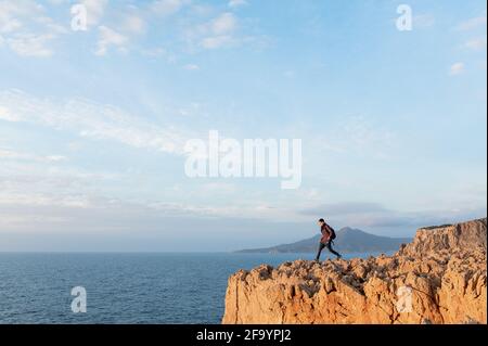 Young hiker man walking on a high jagged cliff overhanging the sea.