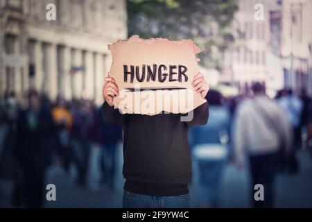 Anonymous man covers his head using a torn cardboard banner with hunger text message. Incognito person on a crowded street as protest demonstration ag Stock Photo
