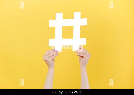 Cropped images of female hands holding large big white hashtag sign, viral web content, internet promotion, isolated on yellow studio background wall Stock Photo