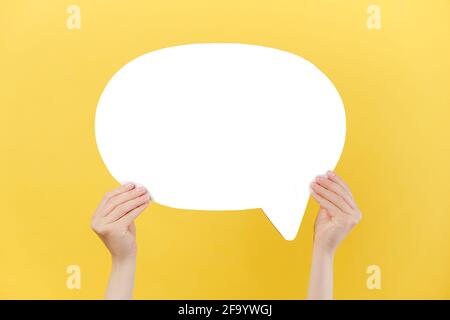 Close up of female hands holding an empty white paper speech bubble, isolated over pastel yellow studio background wall with copy space Stock Photo