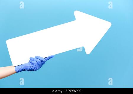 Close up of male in medical protective blue gloves hold white arrow pointing at copy space for advertisement, isolated on blue studio background wall Stock Photo