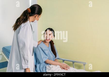Cheerful young Asian patient sitting on hospital bed laughing and smiling while talking to young female doctor in labcoat wearing face mask for safety Stock Photo