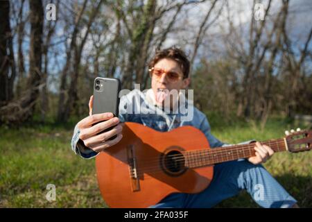Close-up of the hand of a young musician taking a selfie while playing the guitar, he makes a funny face with his tongue out Stock Photo