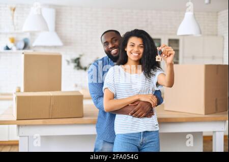 Moving day concept. Happy African-American couple shows keys from new flat standing in embraces surrounded with cardboard boxes, cheerful owners in new property Stock Photo