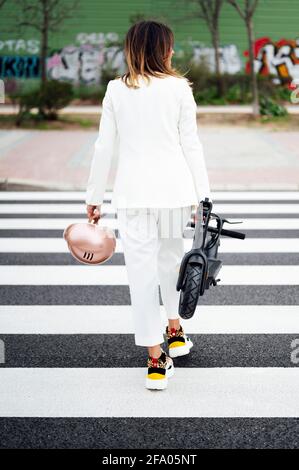 Woman with electric scooter walking on crosswalk seen from behind Stock Photo