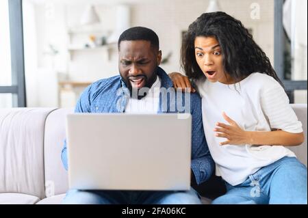 Vexed and surprised African-American couple sits on the couch at home and looks at the laptop screen, received unexpected news, lost in video game Stock Photo