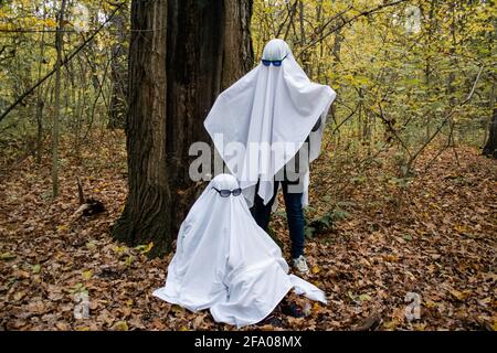 Ghost challenge in the forest or park. Two unrecognizable people teenagers disguised as ghosts with white bed sheets and sunglasses. One stands at ano Stock Photo