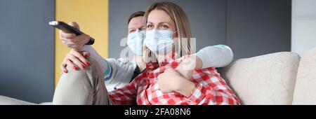 Man and woman in protective medical masks are sitting on couch watching TV Stock Photo