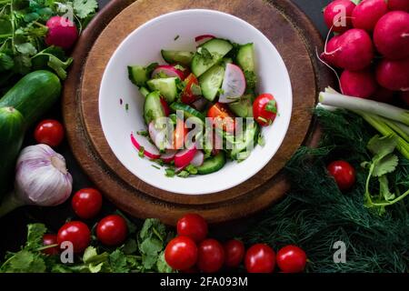 A pile of fresh, ripe vegetables: radishes, tomatoes, cucumbers, herbs, and garlic are arranged around a round wooden chopping board. On it is a white Stock Photo