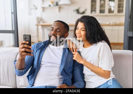 African-American couple with smartphone on the couch spends leisure time in social networks, scrolling feed, watching videos, a black guy is showing smth to his girlfriend Stock Photo