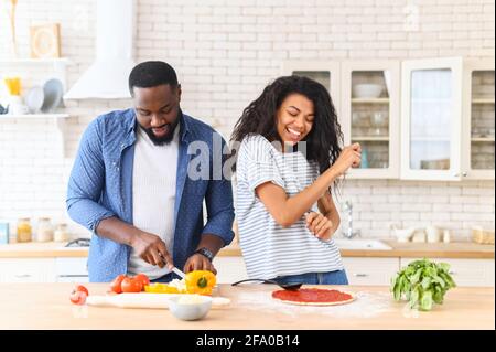 African American couple husband and wife having fun dancing in the kitchen, cooking together at home. Best friends, two people in love preparing cooking pizza, guy chopping veggies, and girl moving Stock Photo