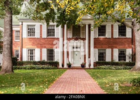 Sidewalk leads to upscale house with white shutters and columns and rocking chairs on porch through tall trees and green grass scattered with yellow l Stock Photo