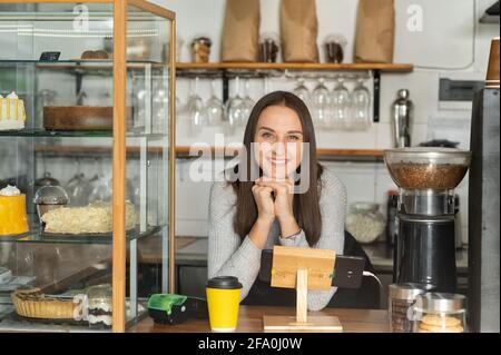 Friendly and smiling waitress in apron leaned on the counter bar and looks at the camera and smiles. Cheerful female bakery owner Stock Photo