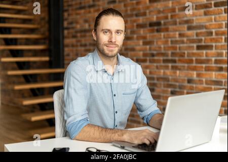 Smiling intelligent male freelancer using laptop for work online sitting at the desk and looks at camera in the coworking space, typing on the keyboard, writes article, chatting and messaging online Stock Photo