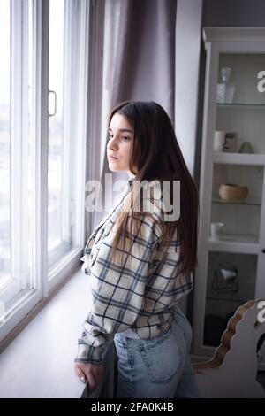 Thoughtful brown haired female student in casual outfit looking away while standing near window at home Stock Photo