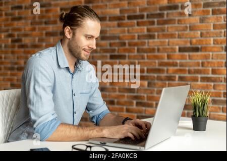 Friendly intelligent male freelancer using laptop for work online sitting at the desk in the coworking space, a thoughtful guy typing on the keyboard, writes article, chatting and messaging online Stock Photo