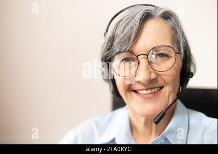 Close-up of the face of a charming beaming elegant elderly lady, wearing headset, glasses, smiling and looking at the camera. Portrait of a senior customer service representative sitting in the office Stock Photo