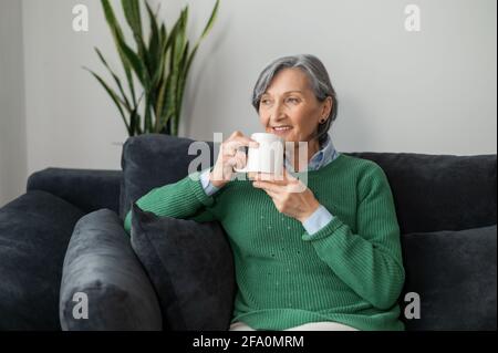Charming senior mature lady is sitting on the sofa, daydreaming and enjoying a hot drink, holding a cup of tea or coffee and looking in the window, grandmother enjoying her morning routine Stock Photo