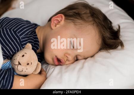 Authentic portrait sick cute caucasian little preschool baby boy in blue sleep with teddy bear on white bed. child resting at lunchtime. care Stock Photo