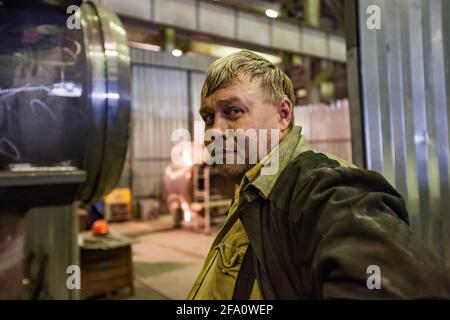 Gas and oil equipment production plant. Young caucasian welder worker in protective jacket posing and looks in camera. Ust-Kamenogorsk, Kazakhstan. Stock Photo