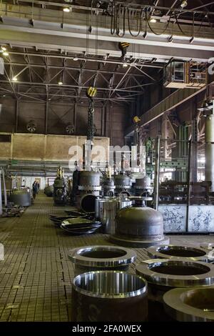 Worker lifts giant shut-off valve by girder crane. Oil and gas industry equipment production plant workshop. Parts of valve on the floor. Stock Photo