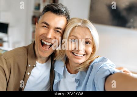 Portrait of beautiful cheerful marriage mature couple in casual clothes, Caucasian husband and wife taking selfie on smartphone, having fun together, looking at camera, smiling Stock Photo