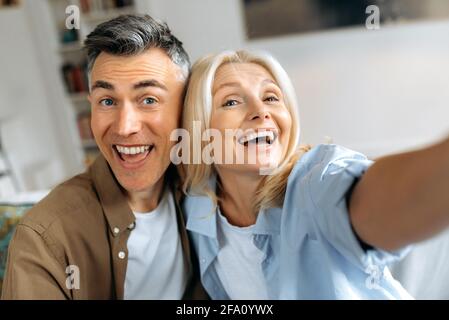 Close-up headshot of happy cute funny mature spouses in casual clothes, Caucasian husband and wife taking selfie on smartphone in their living room, looking at camera, smiling, spend leisure time Stock Photo