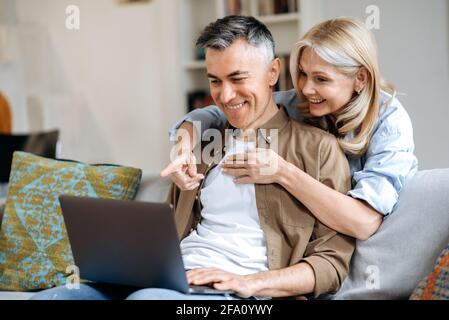 Joyful mature caucasian couple spend time at home, sitting on sofa in casual stylish wear, using laptop for online conference with friends or family, browsing internet, gesturing and smiling friendly Stock Photo