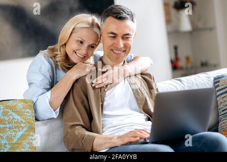 Joyful mature loving caucasian couple at home on sofa in casual stylish clothes, using laptop, browsing social media, chatting with friends or family, shopping online, hugging and smiling Stock Photo