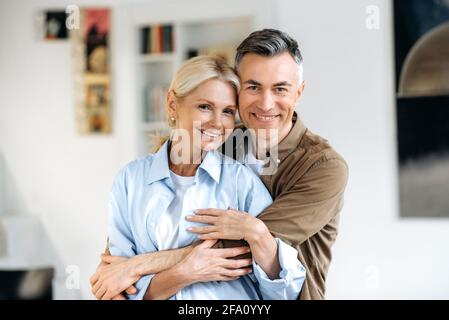 Family portrait. Close-up portrait of joyful happy mature caucasian married couple, stylishly dressed, husband and wife standing in living room, gently hugging each other, looking at camera, smiling Stock Photo