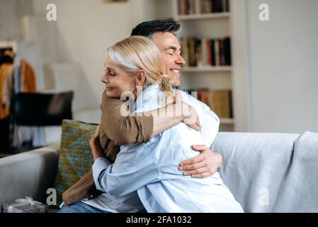 Happy loving senior caucasian family, happy husband and wife dressed in casual stylish clothes, sitting at home on sofa in living room, hugging, eyes closed, spend time together, smiling Stock Photo