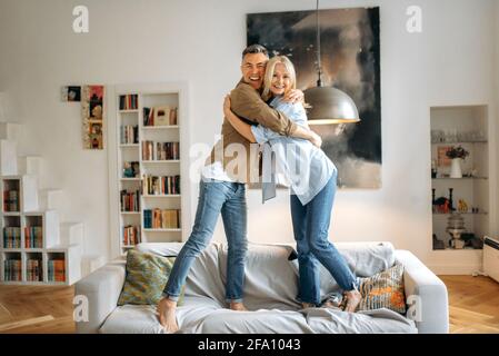 Happy cute joyful mature caucasian married couple dressed in casual stylish clothes, husband and wife are standing on the sofa at home, hugging, smiling, looking at the camera, having fun together Stock Photo