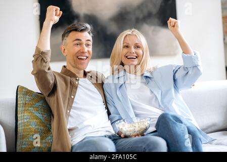 Joyful middle aged Caucasian married couple, husband and wife, sitting at home on sofa in casual stylish clothes, watching TV show, movie or football, eating popcorn, gesturing, rejoicing of winning Stock Photo