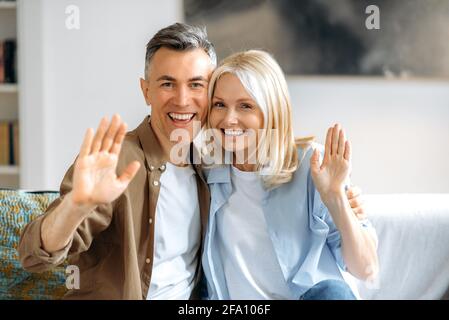 Virtual video meeting. Friendly joyful mature caucasian married couple in stylish casual clothes chatting on video call with friends or family, welcoming hand gesture, looking at camera, smiling Stock Photo
