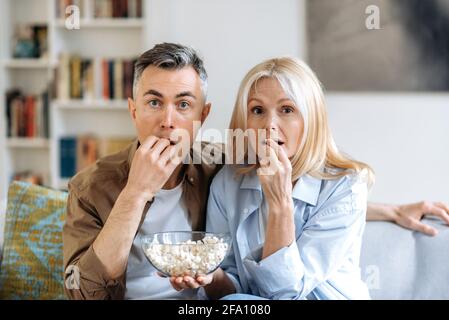 Middle aged shocked caucasian family couple sitting at home on sofa in casual clothes, watching tv show or exciting movie, eating popcorn, spending weekend together at home Stock Photo
